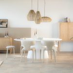 Stijlvolle eetkamer met een witte ovale eettafel van Eleonora, omringd door beige gestoffeerde stoelen en drie hangende rotan lampen. Aan de muur hangt kunst en een moderne klok, terwijl een houten dressoir en kast de ruimte completeren.