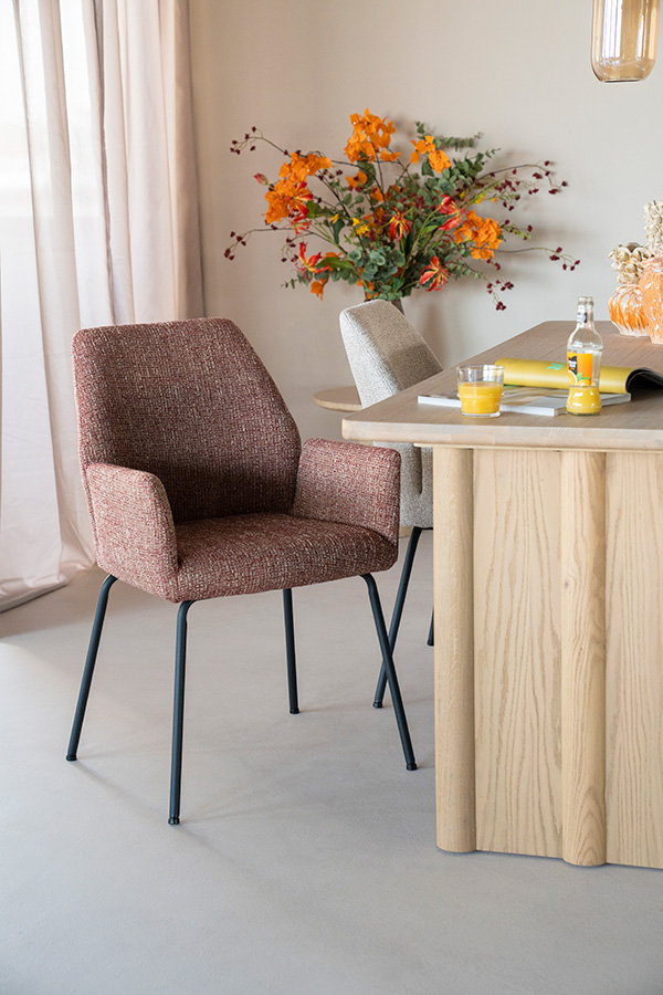 Eetkamerstoel met hoge rug en armleuningen in roodbruine gemêleerde stof aan een lichte houten tafel, met herfstboeket op de achtergrond.