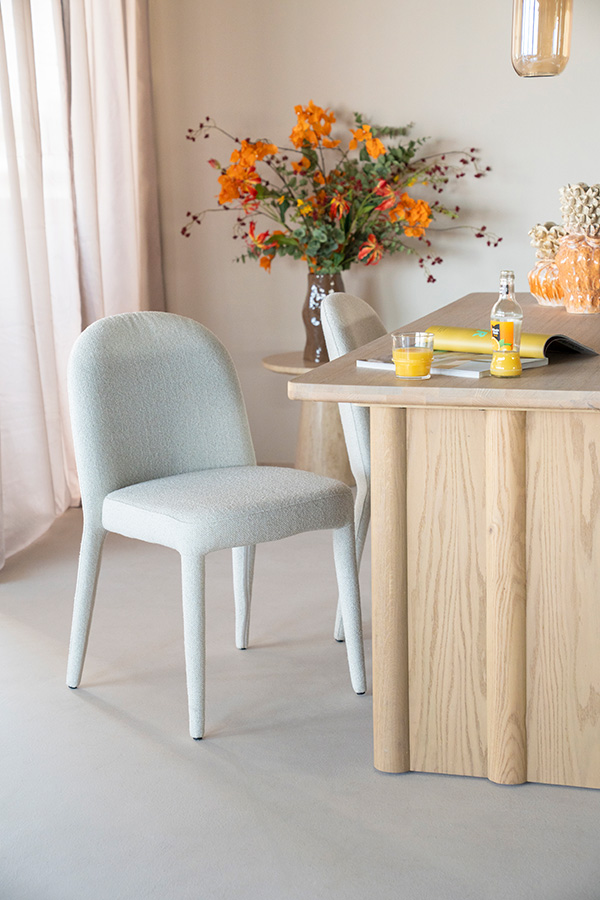 Gestoffeerde eetkamerstoel met afgeronde rugleuning en poten in lichtgrijze stof naast een houten tafel, in een ruimte met zachte natuurlijke styling.