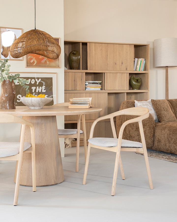 Eettafel Nola en stoel Rayven – naturel Licht eiken eetkamerstoel aan ronde eiken eettafel in lichte woonkamer met lichtgrijze gietvloer.
